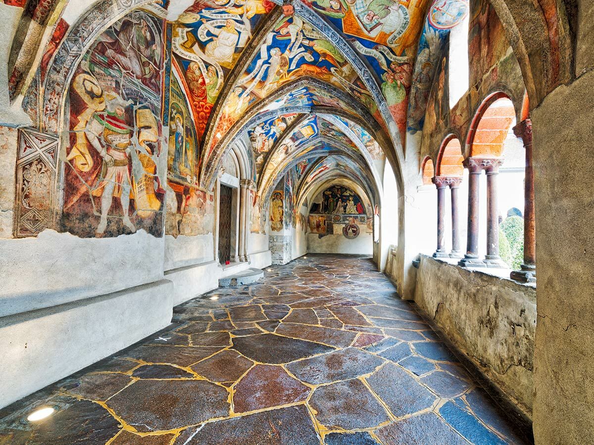Historischer Klostergang mit bunten Fresken an Wänden und gewölbter Decke, Steinfliesenboden und Bogenfenstern zu einem Hof.