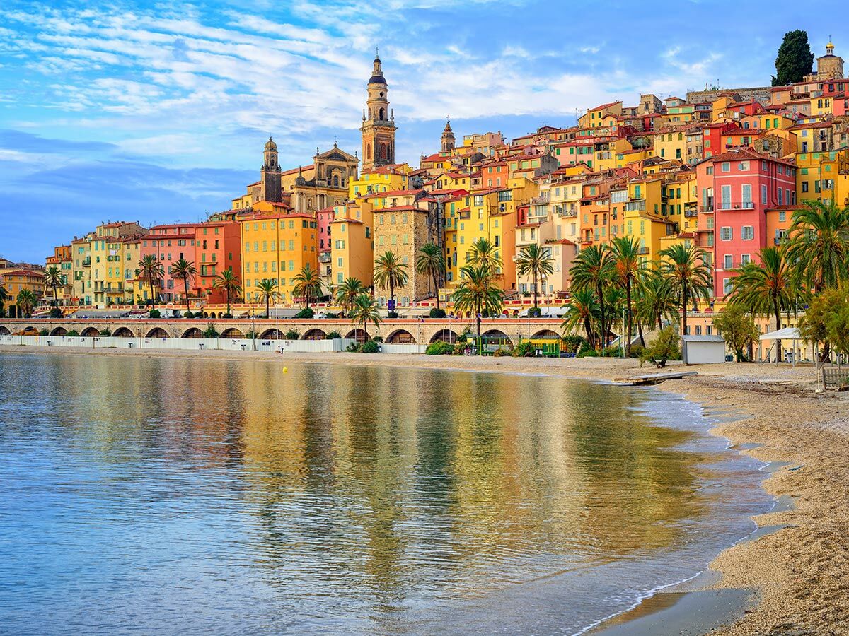 Panoramablick auf die bunten Häuser von Menton, Frankreich, am Mittelmeer mit Strand, Palmen und klarem Wasser.