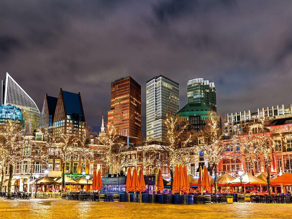 Abendansicht des beleuchteten Stadtzentrums von Den Haag mit modernen Gebäuden, festlichen Lichtern und Outdoor-Cafés.