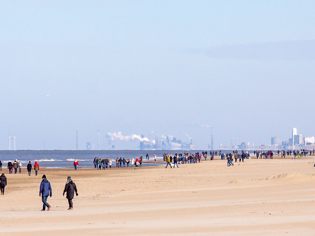 Viele Menschen spazieren an einem breiten Sandstrand mit Meer. Im Hintergrund sind Industrieanlagen, Windräder und eine Stadt sichtbar.