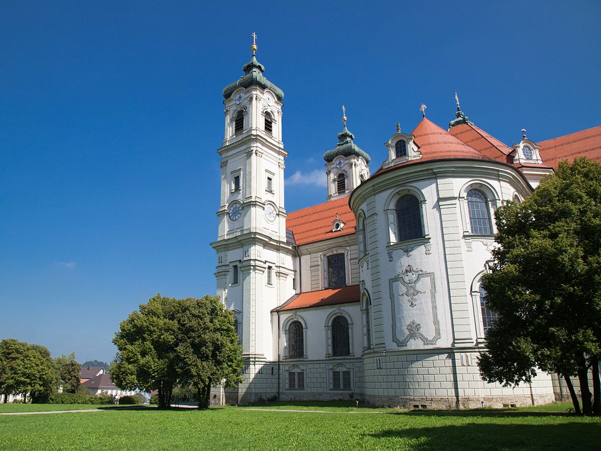 Kloster Andechs an sonnigem Tag mit blauem Himmel, imposanter Barockkirche, Glockenturm und grünen Wiesen.