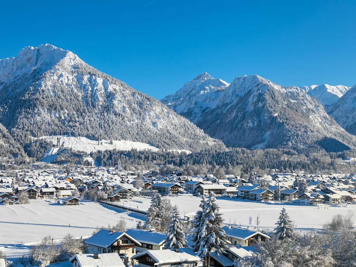 Luftaufnahme eines verschneiten Alpendorfs mit Häusern, Tannenbäumen, majestätischen Bergen und einer Skisprungschanze unter blauem Himmel.