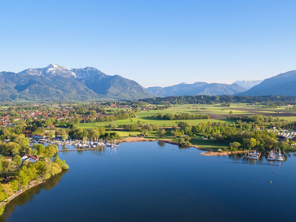 Luftbild eines Alpensees mit Jachthäfen, einem Dorf am Ufer und schneebedeckten Bergen unter blauem Himmel.