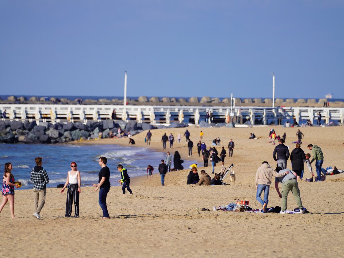 Menschen genießen einen sonnigen Tag am Sandstrand neben einem langen Pier, mit blauem Meer und klarem Himmel im Hintergrund.