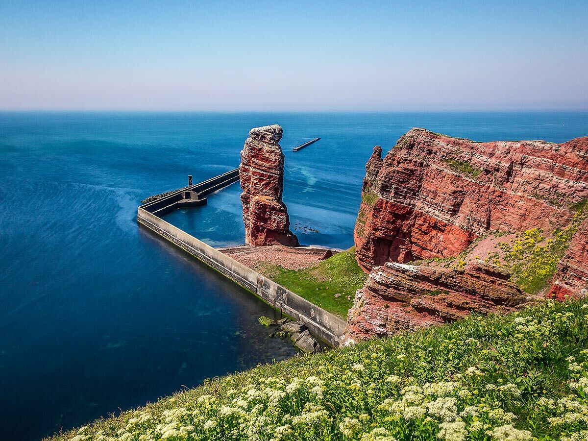Panoramablick auf die Lange Anna, eine markante rote Felsnadel auf Helgoland, umgeben von blauem Meer und grünen Klippen mit Betonmauer.