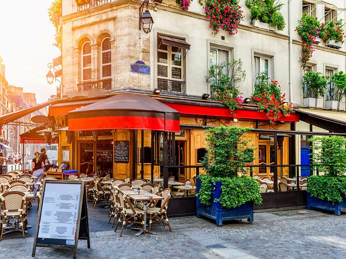Belebtes Pariser Straßencafé mit roten Markisen, vielen Tischen, Stühlen und blühenden Blumen an der historischen Fassade.