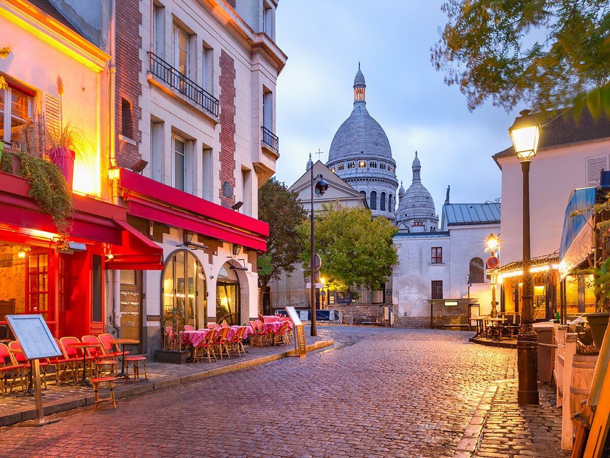 Malerische Kopfsteinpflastergasse in Montmartre, Paris, bei Abenddämmerung mit beleuchteten Cafés und Sacré-Cœur im Hintergrund.