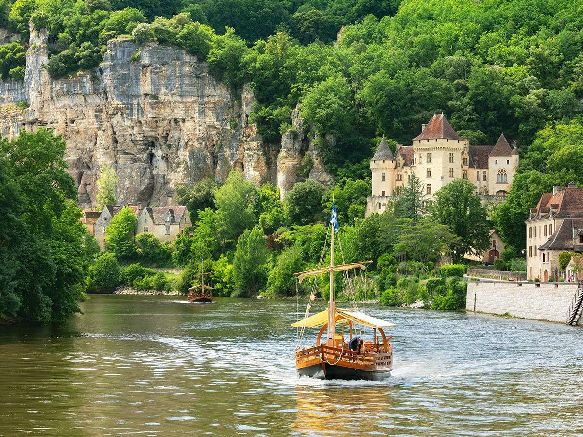 Historische Gabarre auf dem Dordogne Fluss vor einem malerischen Schloss und bewaldeten Klippen in Frankreich.