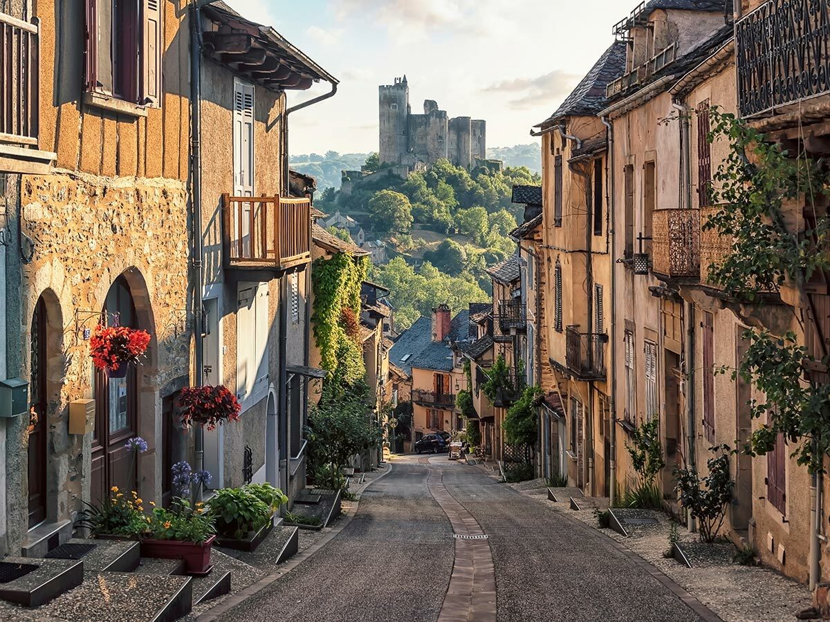 Enge, historische Dorfstraße mit charmanten Häusern, Blumenkästen und einem Blick auf eine imposante Burgruine im Hintergrund.