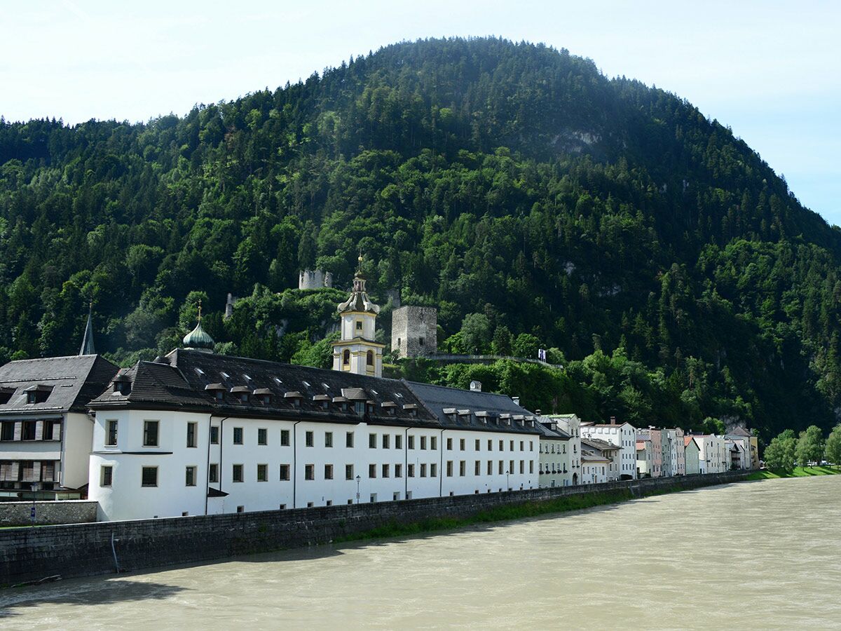 Blick auf St. Johann in Tirol mit historischer Architektur am Fluss und bewaldetem Berg mit Burgruinen im Hintergrund.