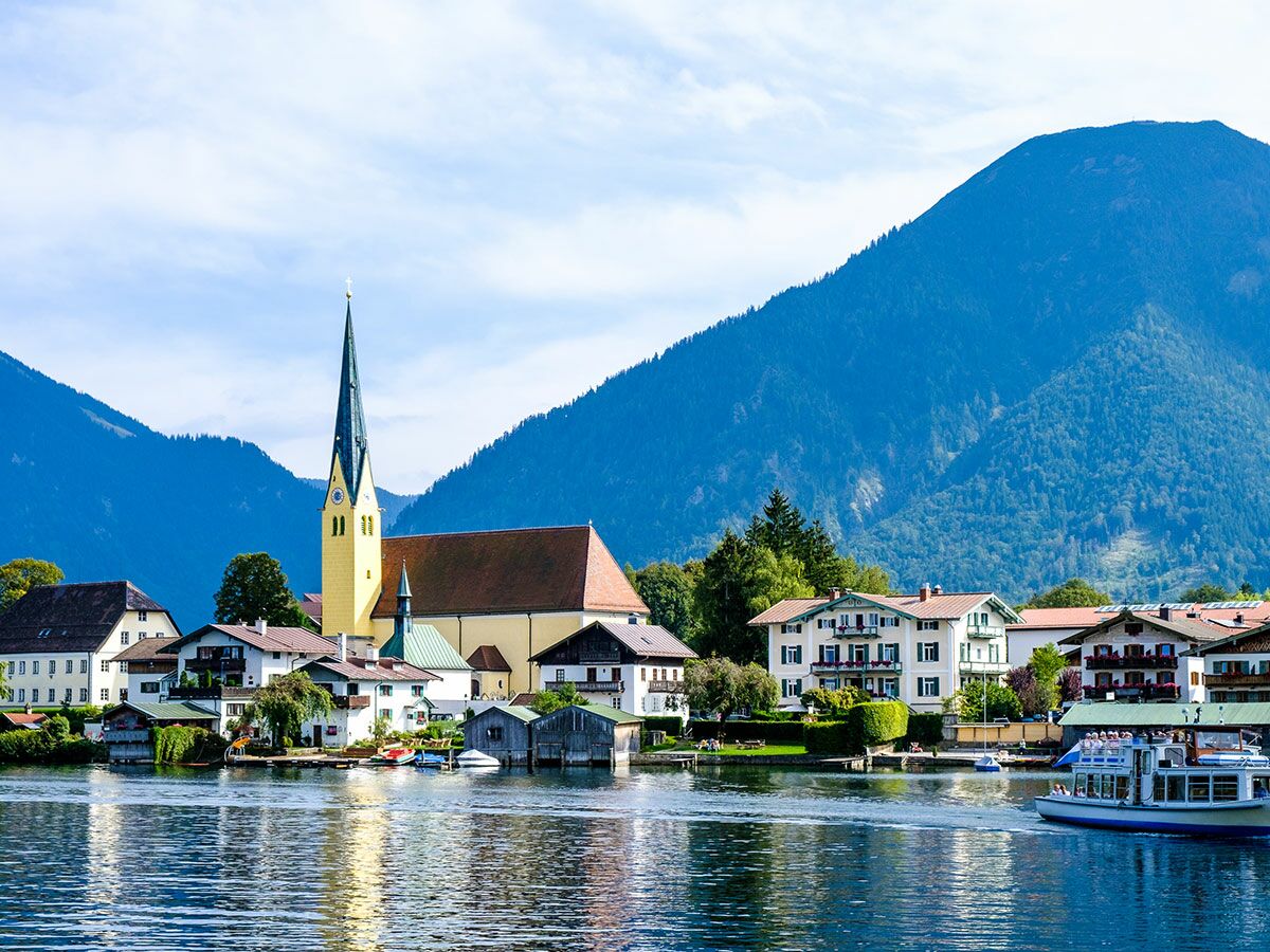 Panoramablick auf idyllisches Dorf am Alpensee mit historischer Kirche, traditionellen Häusern, Bergen und Schiff auf dem Wasser.