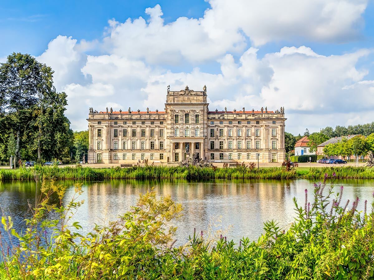 Majestätisches Barockschloss an einem See mit klarer Spiegelung, umgeben von üppiger grüner Vegetation und blauem Himmel mit Wolken.