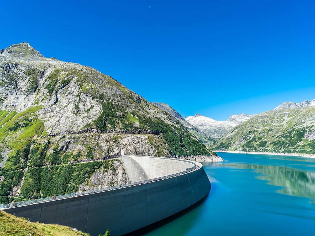Imposanter Staudamm in den Alpen mit türkisblauem Bergsee, umgeben von grünen und schneebedeckten Gipfeln unter blauem Himmel.
