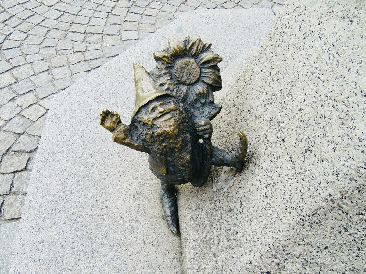Bronze-Zwergfigur mit Spitzhut und Sonnenblume lächelt, ragt aus hellem Granitblock neben Kopfsteinpflaster.