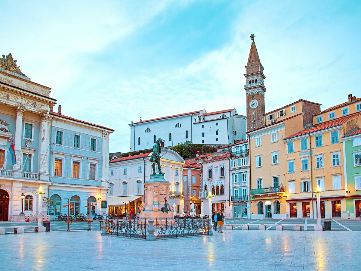 Blick auf den beleuchteten Tartini Platz in Piran, Slowenien, mit Statue, Glockenturm und bunten Gebäuden bei Dämmerung.