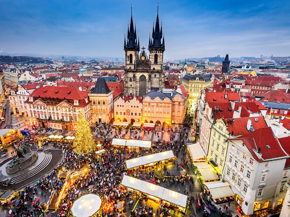 Luftbild des Prager Weihnachtsmarkts auf dem Altstädter Ring mit beleuchteten Ständen, großem Weihnachtsbaum und Teynkirche im Hintergrund.