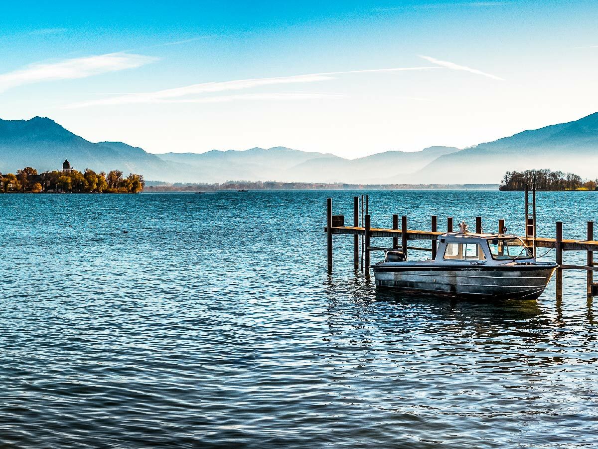 Kleines Motorboot an einem Holzsteg auf einem blauen See, im Hintergrund Berge und eine grüne Insel mit Gebäude.