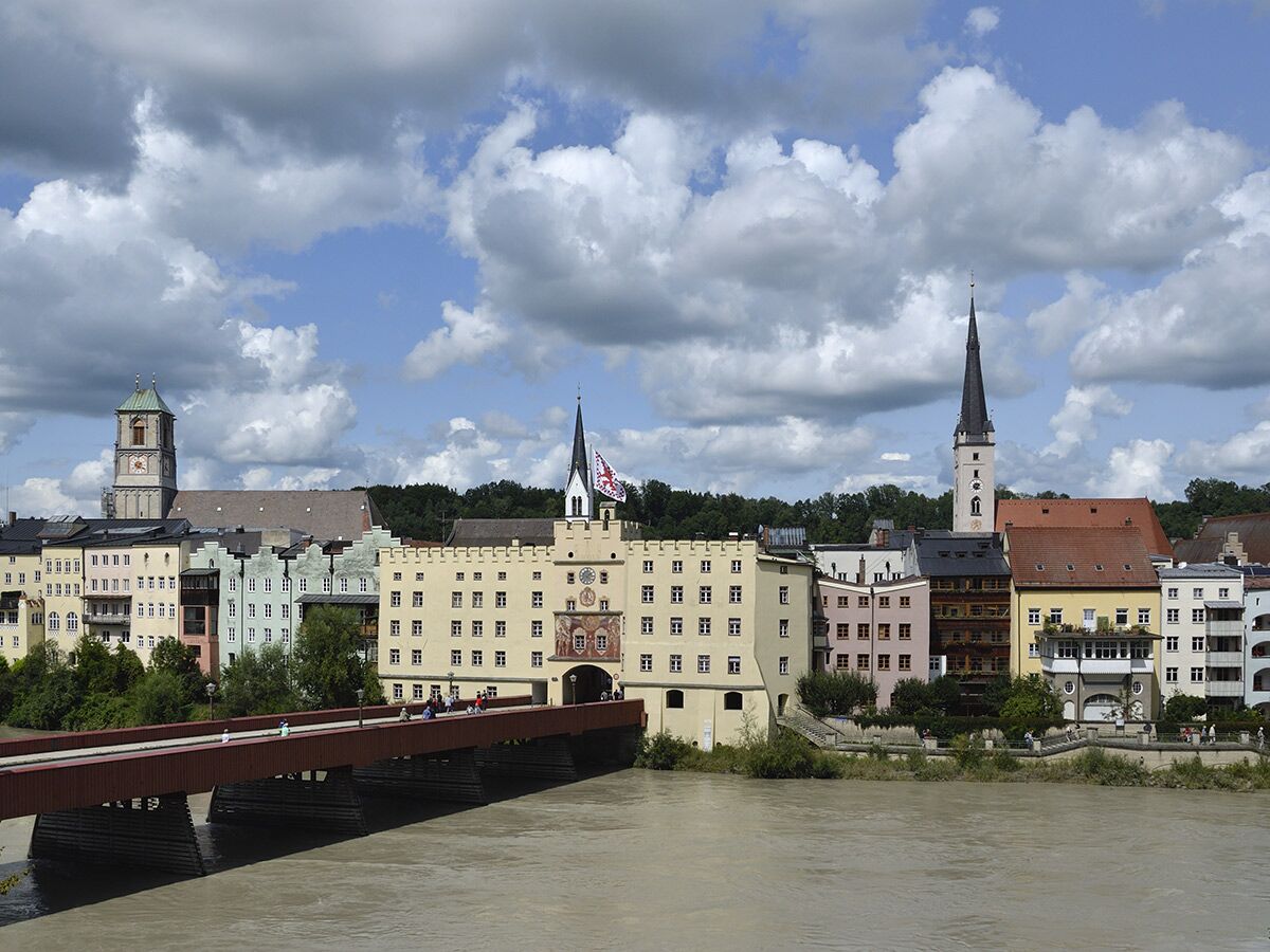 Panoramablick auf eine historische Stadt am Fluss mit farbigen Gebäuden, Kirchtürmen und einer roten Brücke unter bewölktem Himmel.
