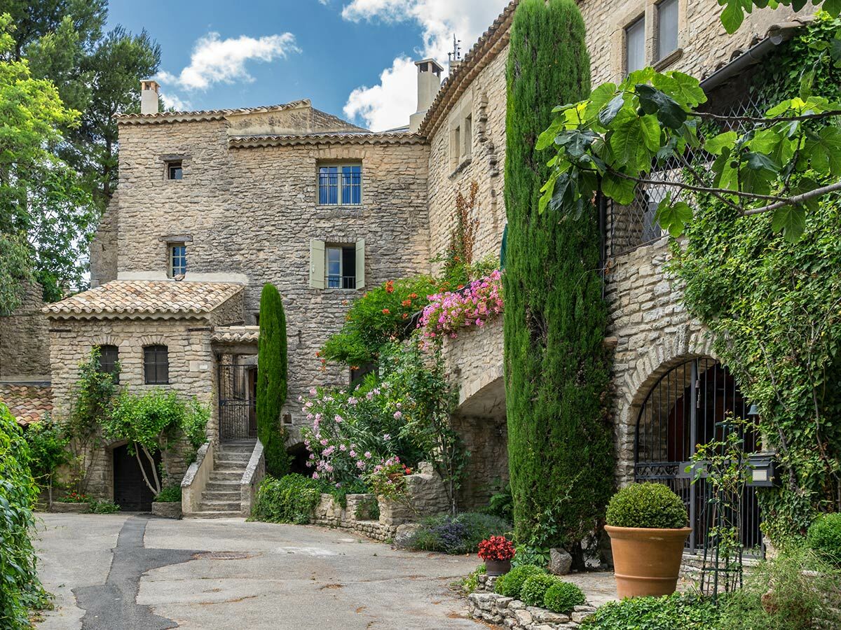 Malerisches Steindorf in der Provence mit historischen Häusern, grünen Pflanzen, bunten Blumen und Zypressen unter blauem Himmel.