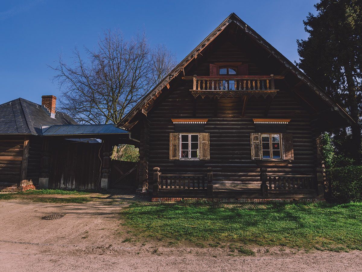 Dunkles, traditionelles Holzhaus mit geschnitztem Balkon, Fensterläden und Verbindungsbau; blauer Himmel, grüne Wiese.