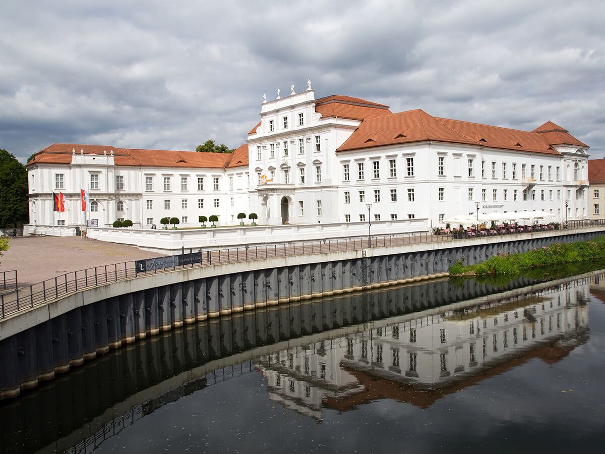 Historisches weißes Schloss mit rotem Dach am Fluss, reflektiert im Wasser, unter bewölktem Himmel. Restaurantterrasse sichtbar.