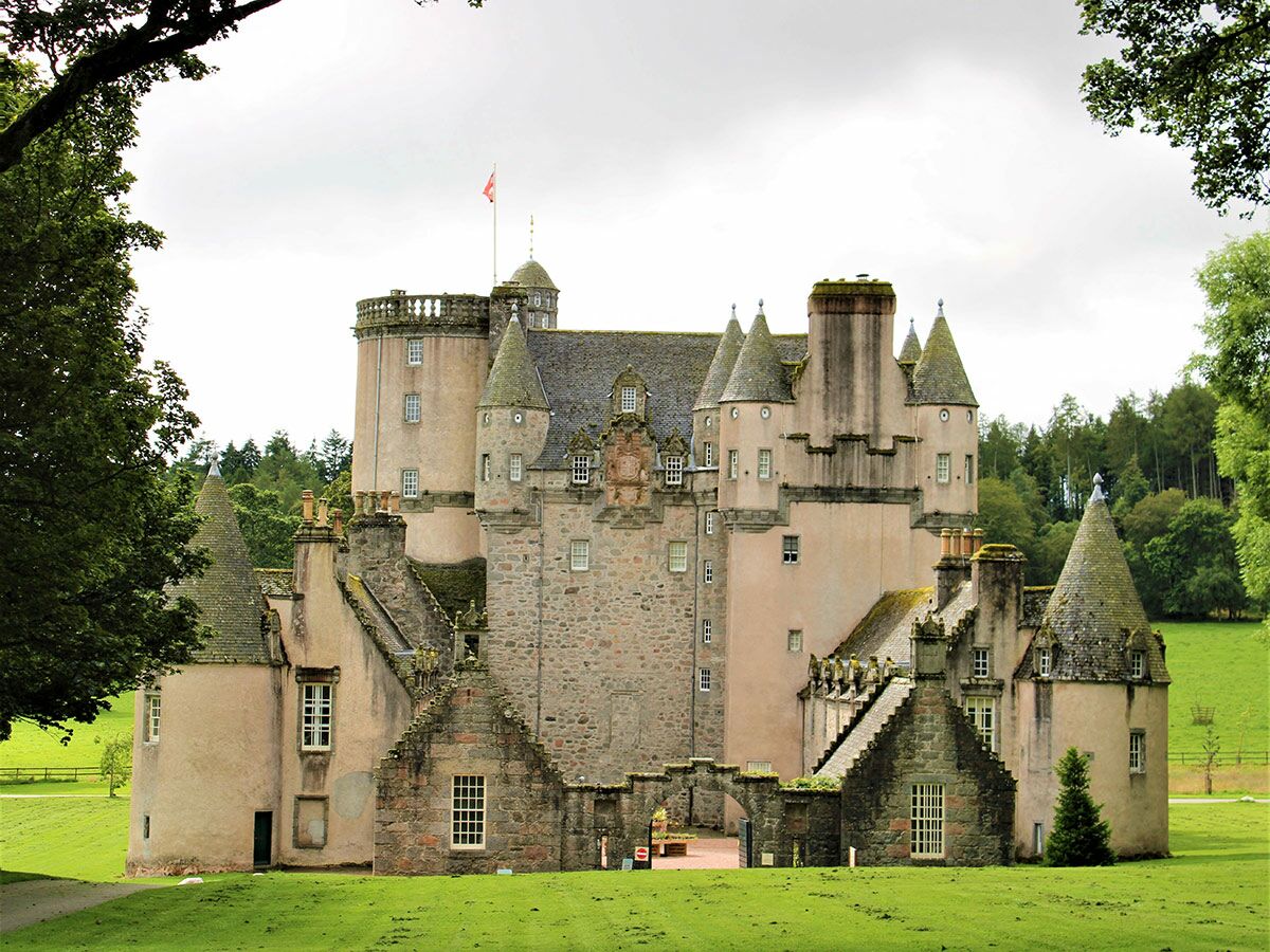 Majestätisches schottisches Schloss mit zahlreichen Türmen und Spitzdächern, umgeben von grünen Wiesen und Wäldern unter bewölktem Himmel.
