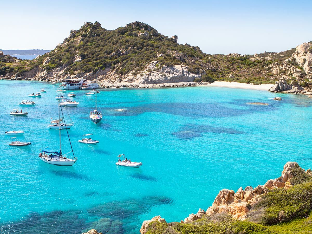 Luftaufnahme einer atemberaubenden türkisen Bucht in Sardinien mit zahlreichen Segel- und Motorbooten am weißen Sandstrand.