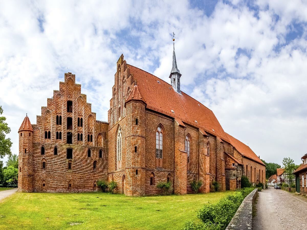 Blick auf ein rotes Backsteinkloster mit Staffelgiebeln und spitzem Turm unter blauem Himmel, umgeben von grünem Rasen.