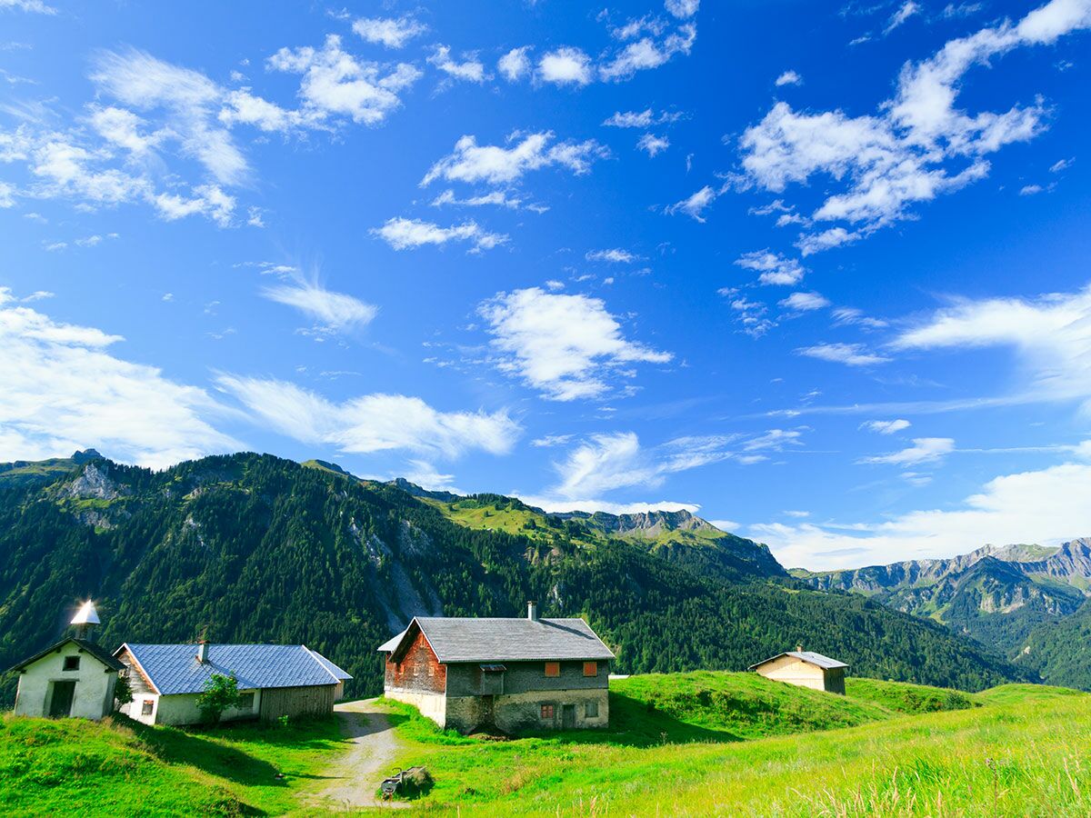 Idyllische Alpenlandschaft mit grünen Wiesen, bewaldeten Bergen, blauem Himmel und traditionellen Almhütten.