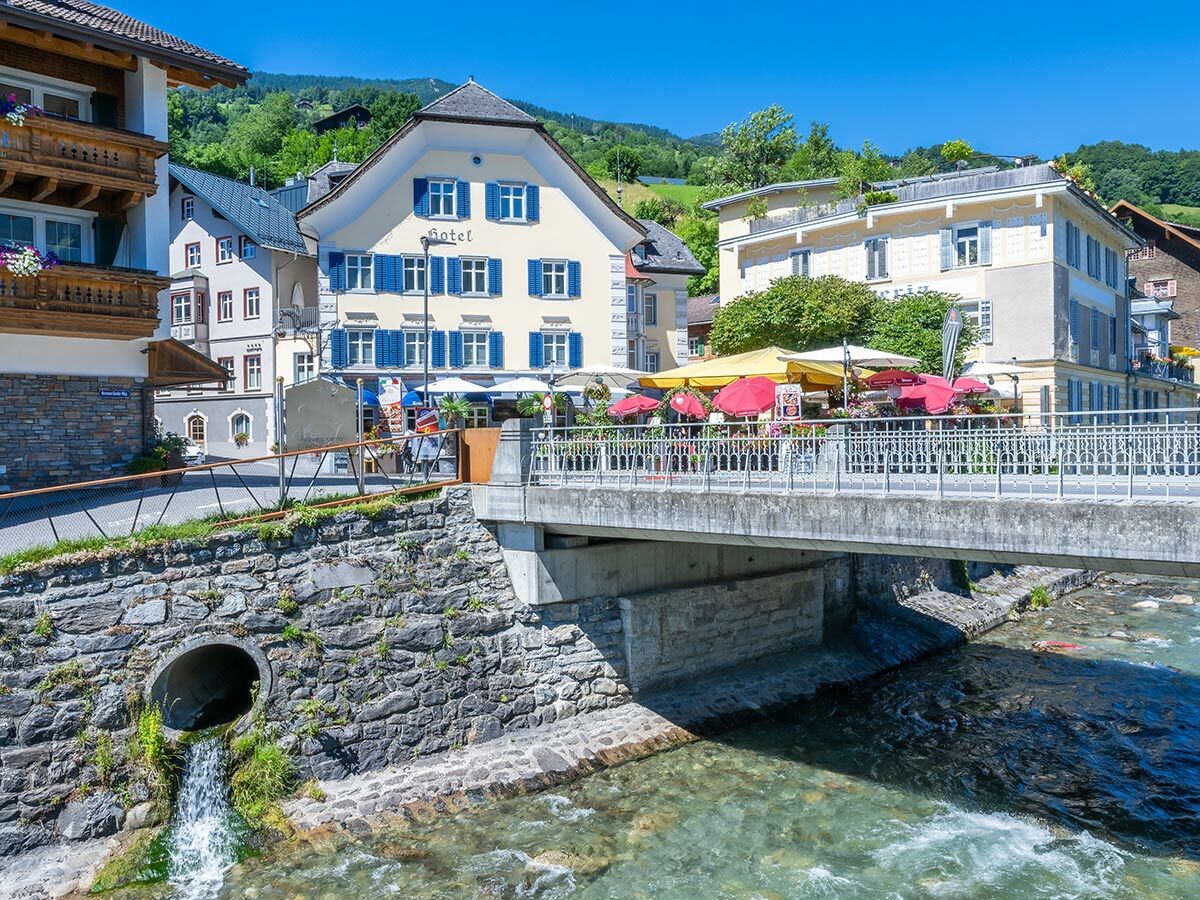 Malerisches Hotelgebäude mit blauen Fensterläden und Außenterrasse an einem Fluss in einem sonnigen Bergdorf.