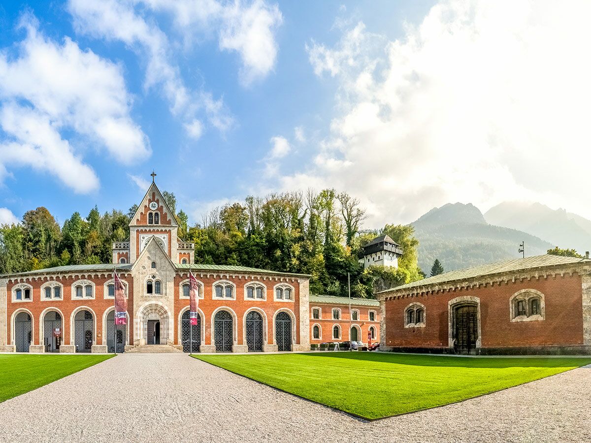 Eindrucksvolle historische Schlossanlage aus rotem Backstein mit zentralem Uhrturm, grünen Wiesen und Alpenhügeln unter blauem Himmel.