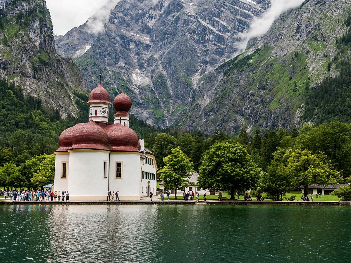 Kirche St. Bartholomä am Königssee, Bayern, mit roten Zwiebeltürmen vor hohen Bergen und grünen Wäldern. Touristen am Ufer.