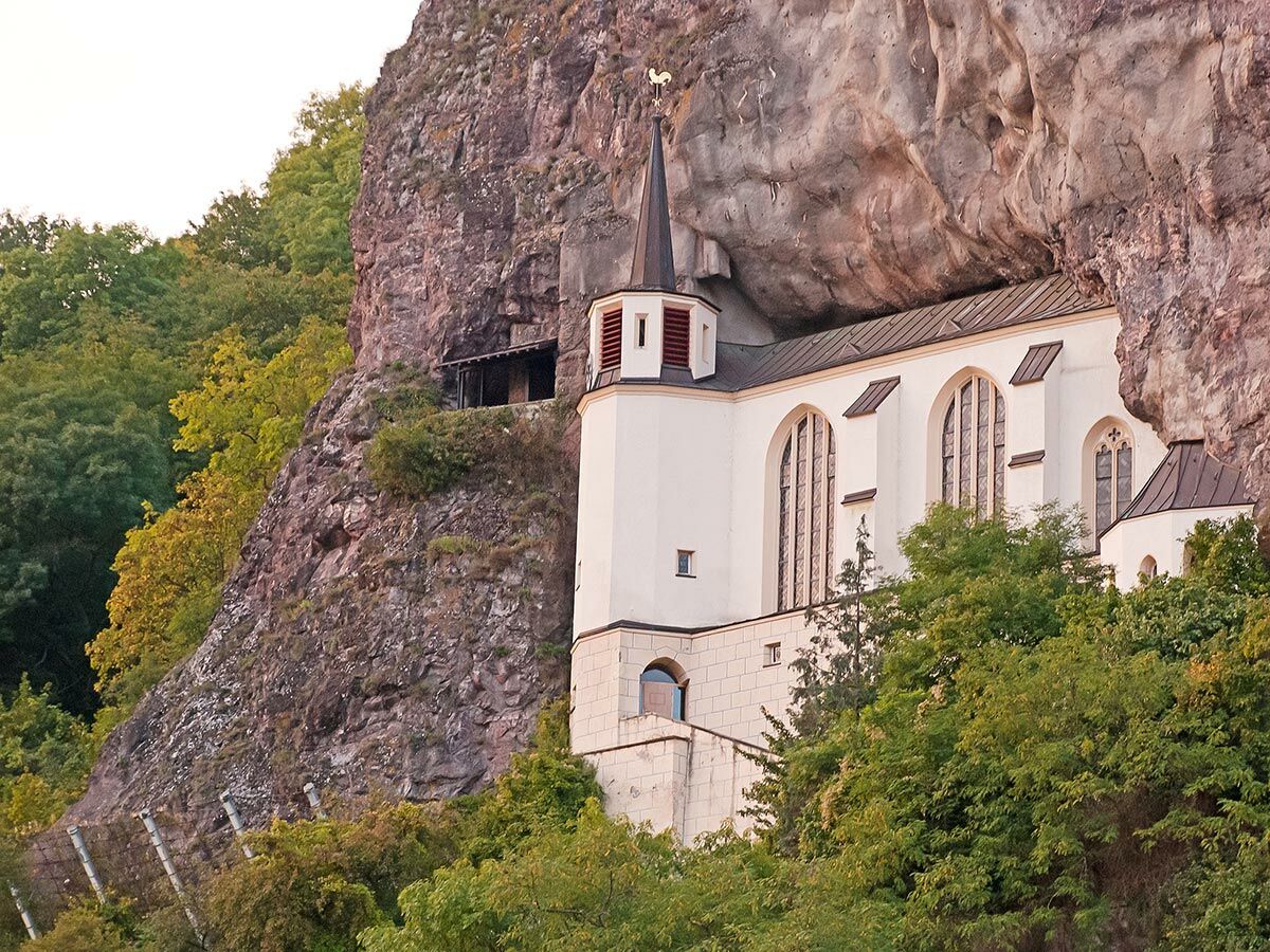 Weiße Felsenkirche in steilem Felsen. Gotische Fenster, spitzer Turm, daneben Bäume und obere Felsenstruktur, unten Schutznetz.