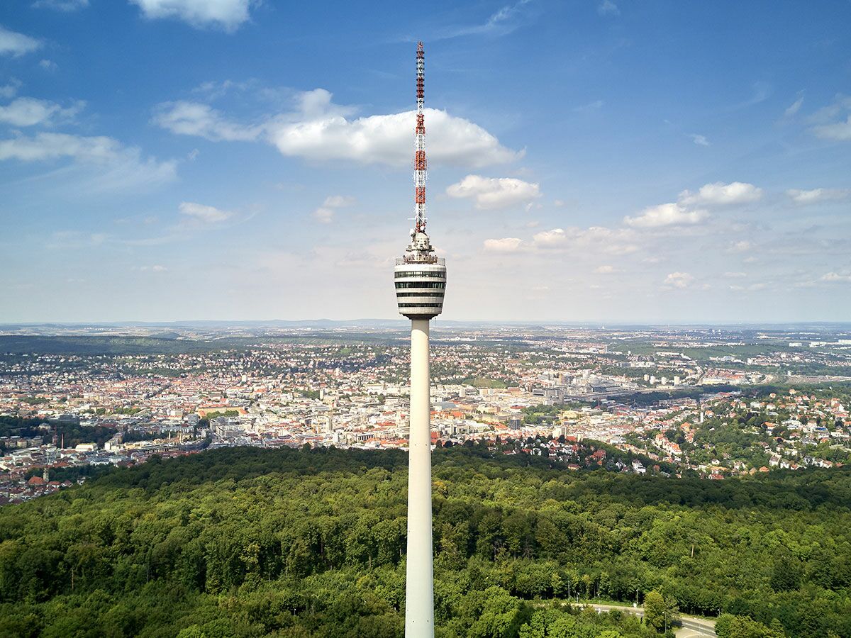 Der Stuttgarter Fernsehturm ragt über die Stadtlandschaft mit Wäldern und Häusern unter blauem Himmel.