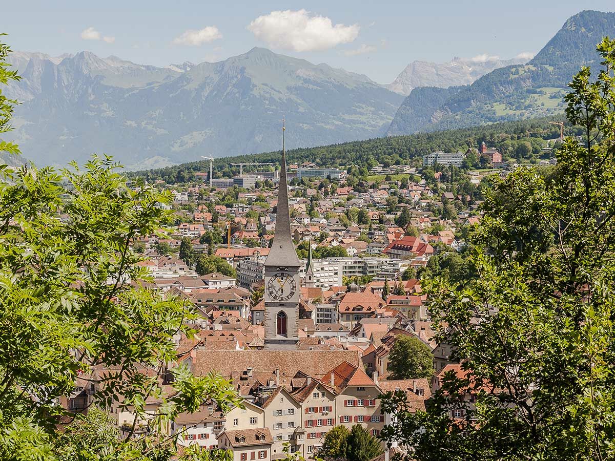 Panoramablick auf eine Alpenstadt mit rotdächigen Häusern, zentralem Kirchturm und majestätischen Bergen, eingerahmt von grünen Bäumen.