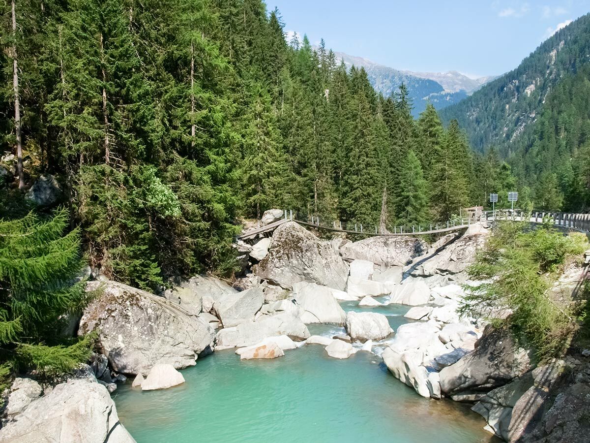Blick auf eine Hängebrücke über einen türkisen Fluss mit Felsen im Gebirge, umgeben von Nadelwald und Bergen.