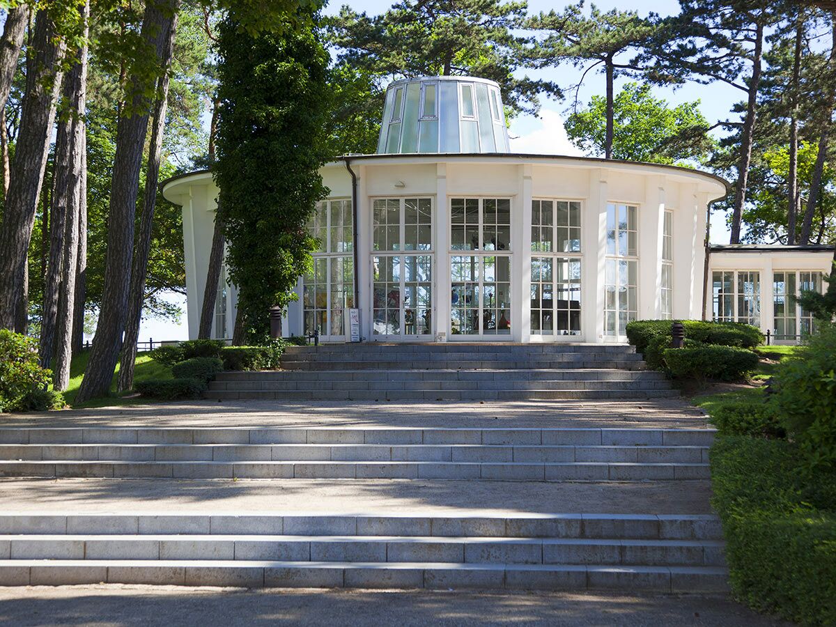Runder, weißer Pavillon mit Glaskuppel und großen Fenstern, umgeben von Bäumen und Büschen, mit breiter Steintreppe.