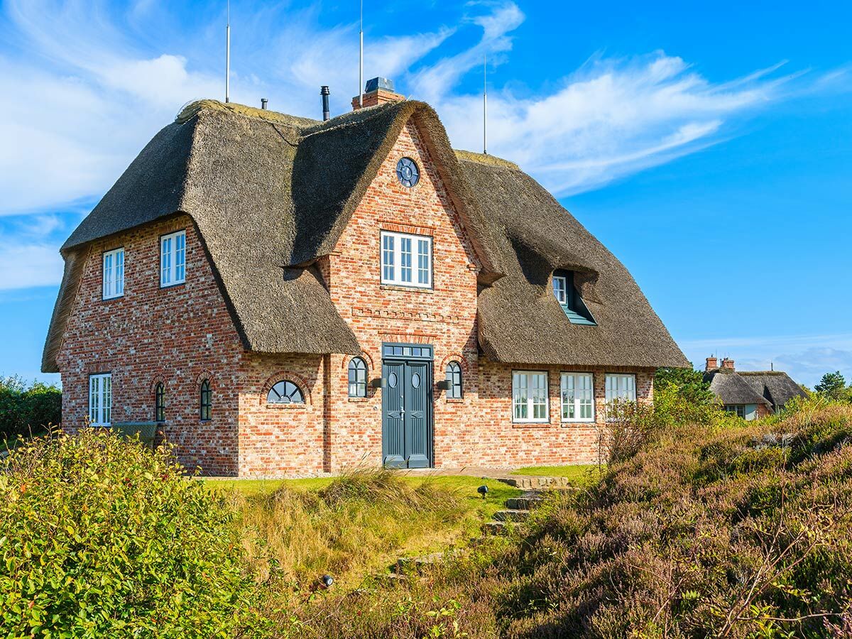 Charmantes reetgedecktes Backsteinhaus mit blauem Himmel und weißen Wolken, umgeben von grünen Büschen und lila Heide.