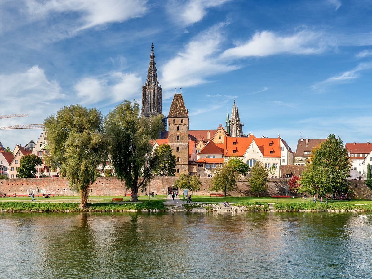 Blick über die Donau auf die Stadt Ulm mit dem Ulmer Münster, dem Metzgerturm und einem grünen Park am Flussufer bei Sonnenschein.