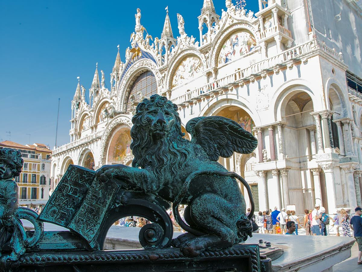 Bronze-Statue des geflügelten Markuslöwen mit Buch, im Hintergrund der detailreiche Markusdom in Venedig unter blauem Himmel. Touristen sind sichtbar.