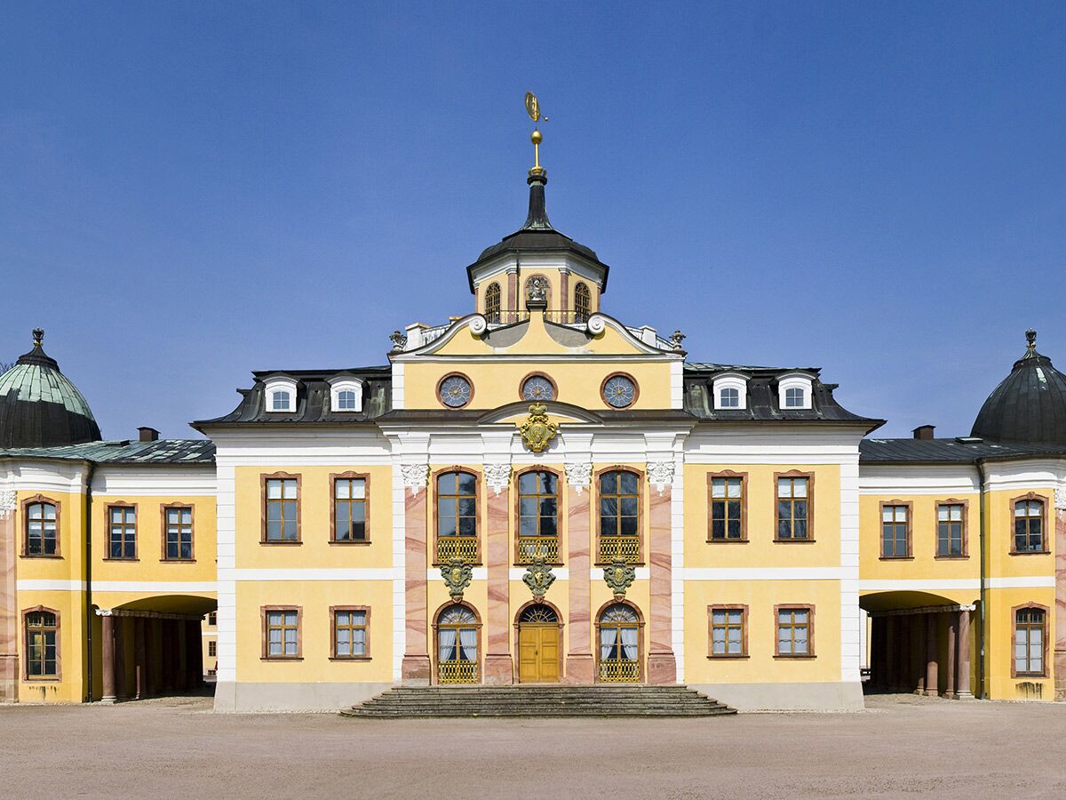 Gelbes Barockschloss mit schwarzen Kuppeldächern und einem zentralen Turm unter strahlend blauem Himmel, detailreich verziert.