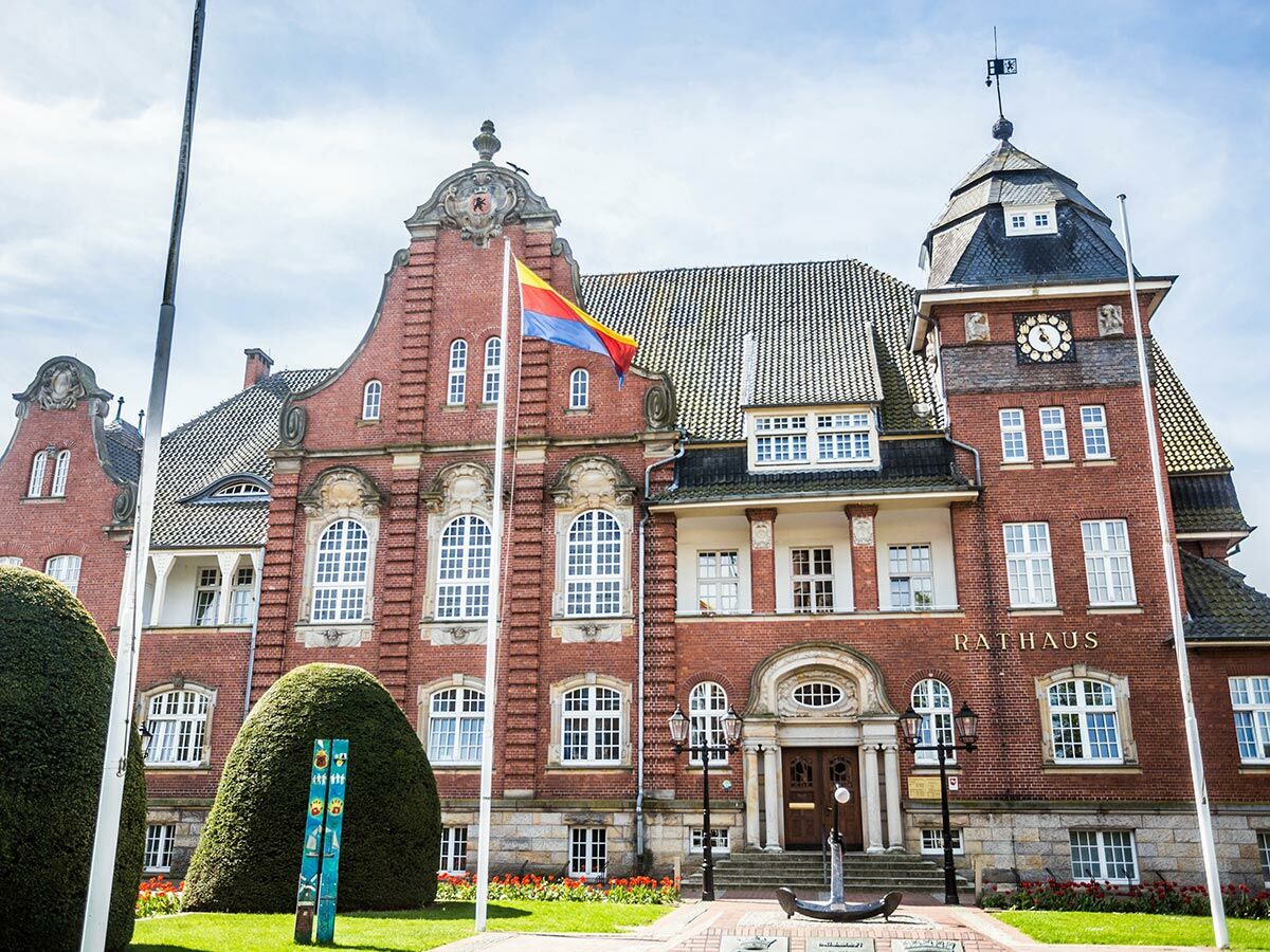 Historisches Backstein-Rathaus mit markantem Turm, Uhr und wehender Flagge vor blauem Himmel. Gepflegte Grünflächen und Eingangsbereich.