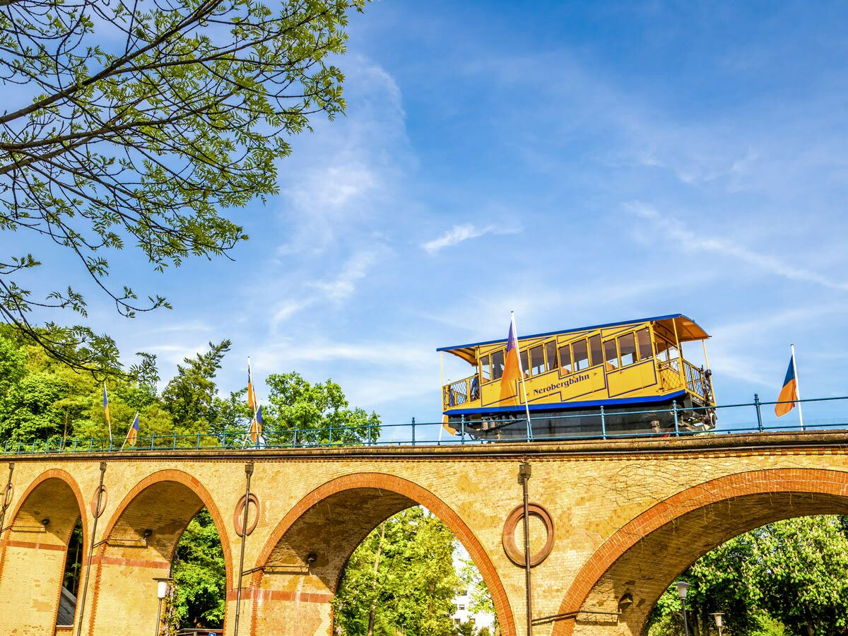 Gelbe Nerobergbahn fährt auf einem alten Steinviadukt mit Bögen, umgeben von grünen Bäumen unter blauem Himmel.