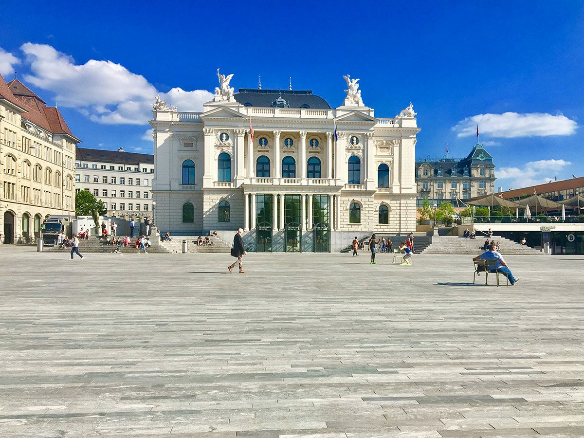 Das imposante Opernhaus Zürich an einem sonnigen Tag mit Menschen auf dem belebten Sechseläutenplatz.