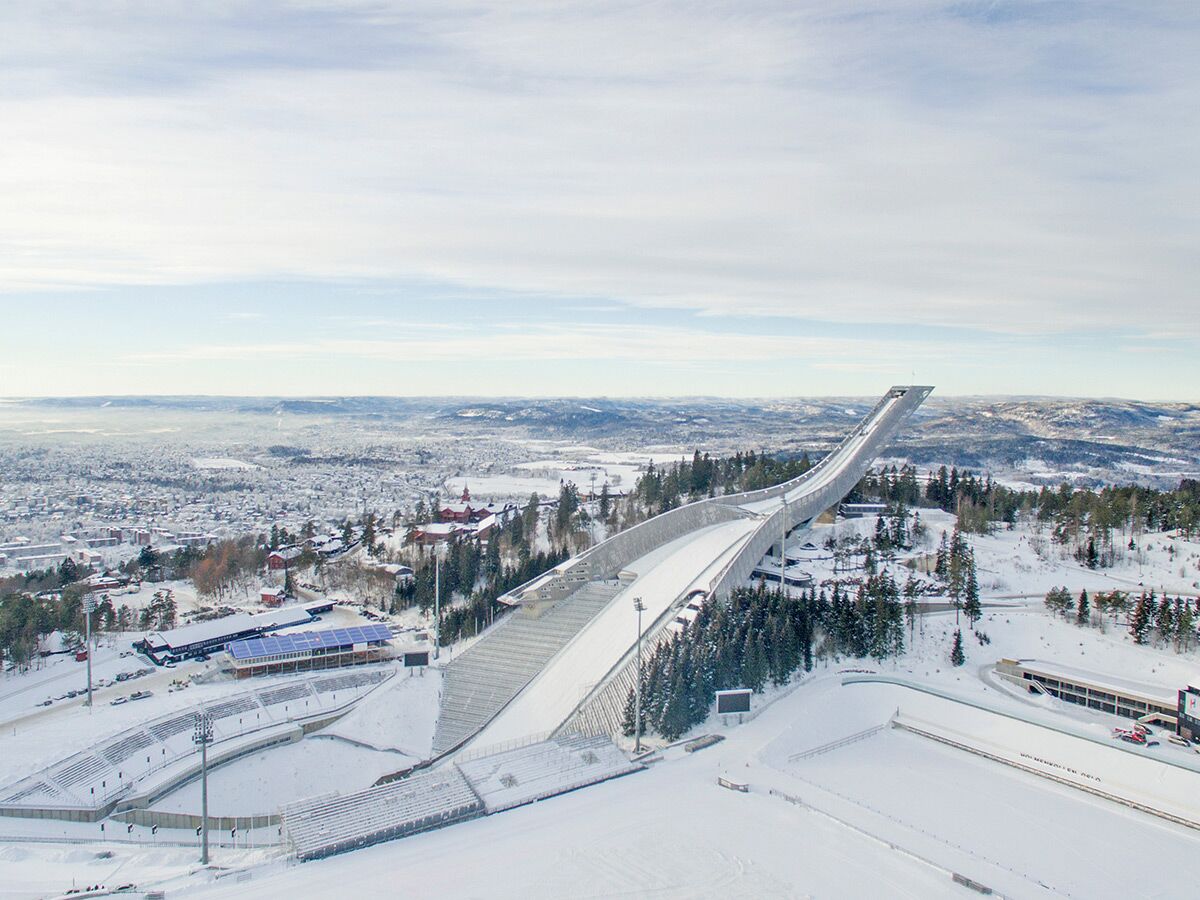 Winterliche Luftaufnahme der Holmenkollen Skisprungschanze in Oslo, Norwegen, mit schneebedeckter Landschaft und Stadt im Hintergrund.
