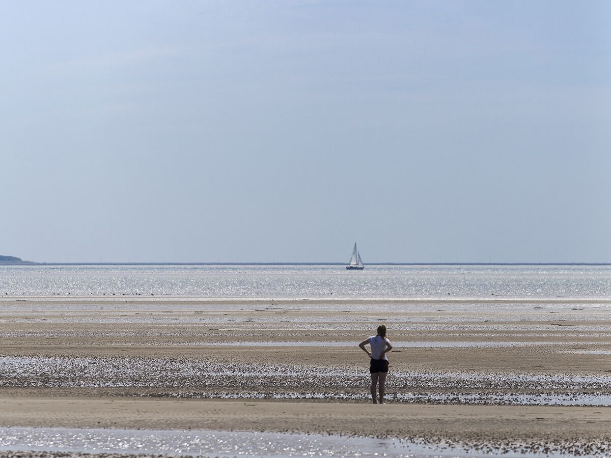 Frau steht am weiten Sandstrand bei Ebbe, blickt auf ein Segelboot auf dem schimmernden Meer unter blauem Himmel.