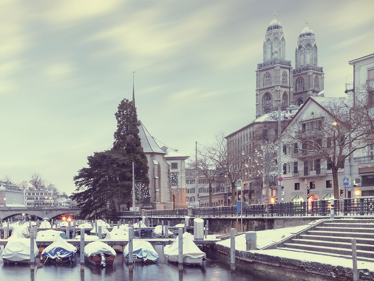 Winterliches Zürich: Schneebedeckte Boote im Fluss Limmat, Grossmünster und Wasserkirche unter bewölktem Himmel.