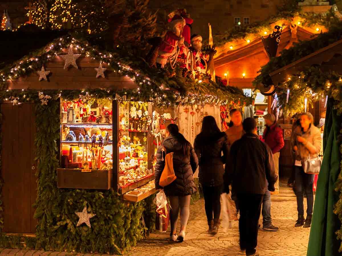 Festlicher Weihnachtsmarkt bei Nacht mit beleuchteten Holzbuden, Girlanden, Sternen und Menschen, die zwischen den Ständen schlendern.