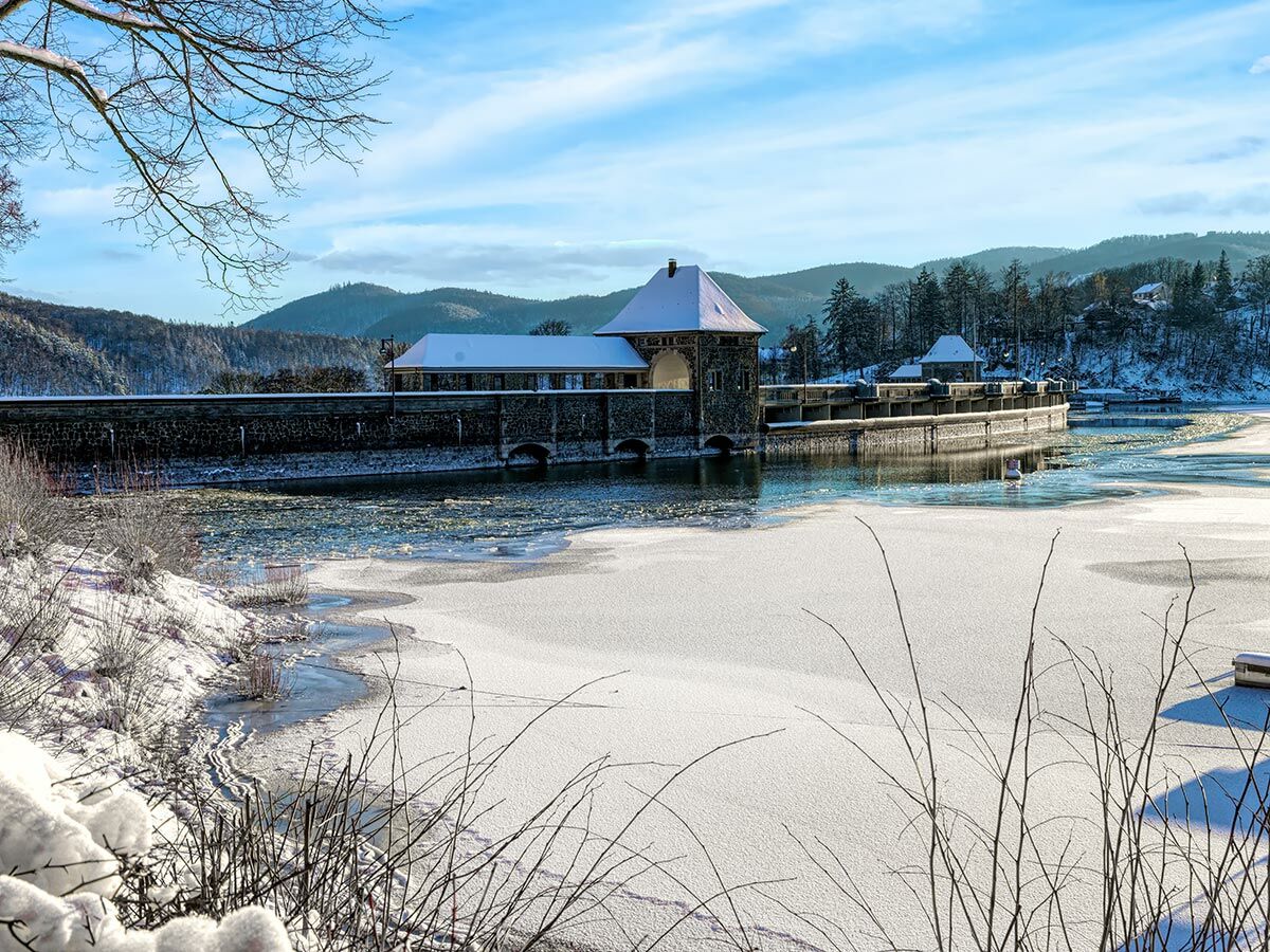 Winterliche Landschaft mit schneebedeckter Staumauer, zugefrorenem See, schneebedeckten Bäumen und Bergen unter blauem Himmel.