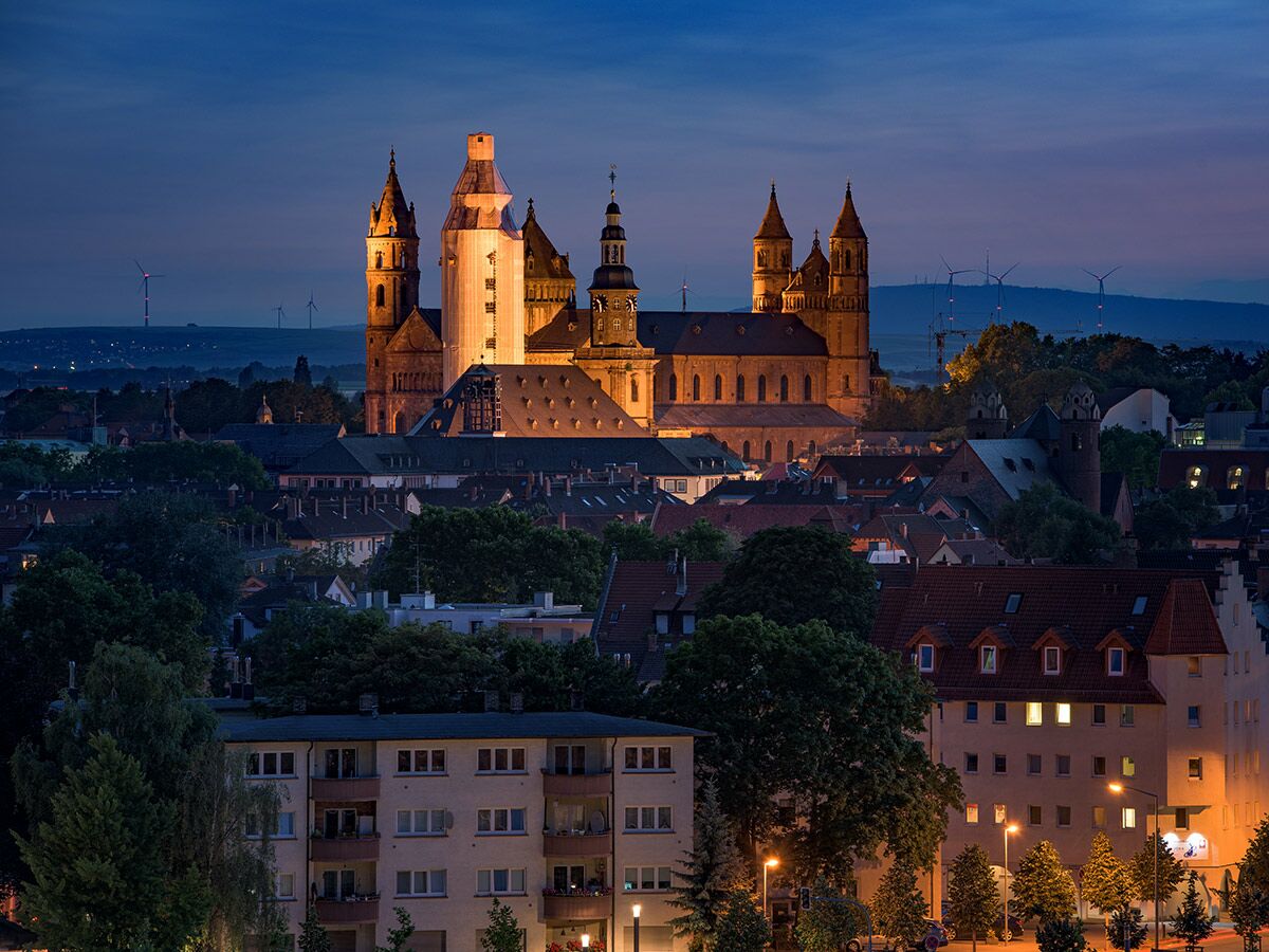 Panoramablick auf den beleuchteten Dom zu Worms und die Altstadt bei Dämmerung mit Windrädern im Hintergrund.
